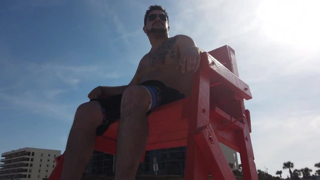 A dolly rotation around a male lifeguard on a high seat at the ocean. Shot at 60fps for optional slow motion usage.  	