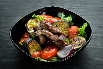 Baked beef and grilled vegetables in a black plate. On a wooden background. Top view. Free space for your text.