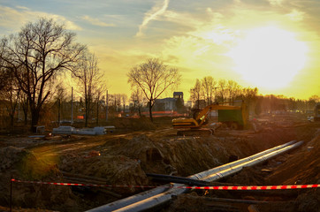 Tracked excavator with a bucket in the ground at a construction site during the laying of sewer pipes in the ground, against a blue sky and sunset. Engineering and heating systems