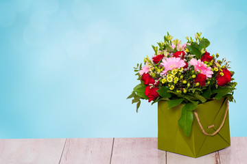 Closeup of a beautiful bouquet with colorful flowers in a decorative green gift box on a bright wooden table over abstract spring blue background. Mothers day, valentine or other festivals.