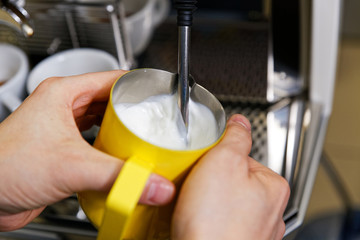 Closeup hands of barista who steaming milk for cappuccino or latte in yellow pitcher.