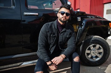 Plakat Fashion rich beard Arab man wear on black jeans jacket and sunglasses posed against big black suv car. Stylish, succesful and fashionable arabian model guy.