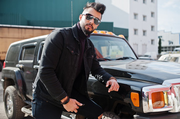 Fototapeta na wymiar Fashion rich beard Arab man wear on black jeans jacket and sunglasses posed against big black suv car. Stylish, succesful and fashionable arabian model guy.