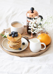 Breakfast in bed. Pancakes with blueberries and honey, coffee, milk and fresh orange served on wooden tray on white sheets.
