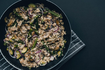 Brown rice with asparagus, courgette, mushrooms and red onion in a pan on stripy grey tea towel on black background with space of copy or text.