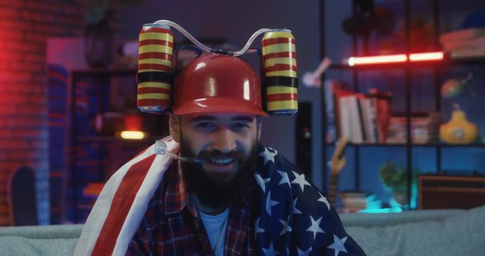 Portrait shot of the young Caucasian handsome guy with a beard sitting on the couch with the American flag and in beer helmet and smiling to the camera..