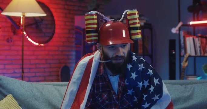 Young Caucasian handsome guy with a beard sitting on the sofa at night with the American flag and in beer helmet and changing channels while watching TV.