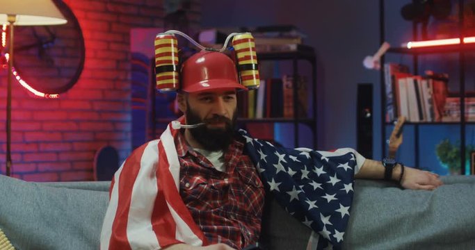 Funny Caucasian young guy sitting on the sofa at home in the evening with the American flag and in beer helmet and changing channels while watching TV.