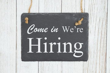 Come in we're hiring text on a hanging chalkboard on weathered whitewash textured wood