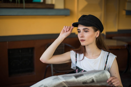 Young woman having a breakfast reading newspaper outdoors at the typical french cafe terrace in France. Playful girl with long hair posing outdoor. Wearing wool cap. Street fashion look. Cute teen