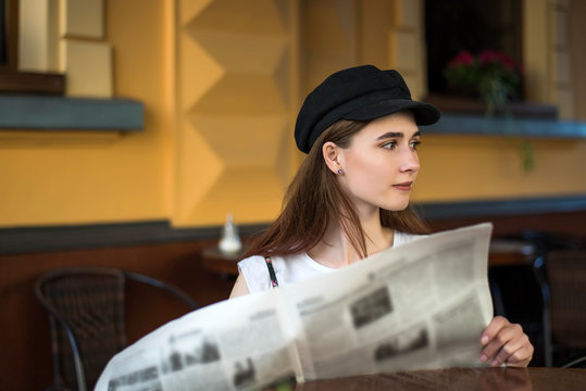 Young woman having a breakfast reading newspaper outdoors at the typical french cafe terrace in France. Playful girl with long hair posing outdoor. Wearing wool cap. Street fashion look. Cute teen