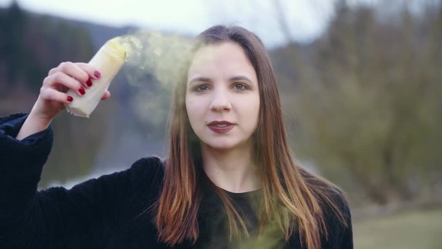 Girl wave with yellow smoke 4K. Static slow-motion shot of a female person in focus looking into the camera and holding a smoke bomb in one hand. Background lake and trees.