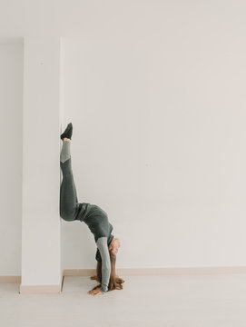 Young sportive woman training upside down in light room studio wall