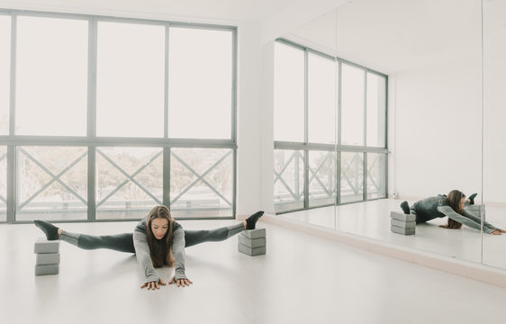 Young slim sportive woman doing leg split near big mirrors in light room