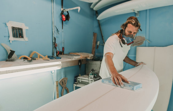 Male in breather with sand paper polishing wooden surf board in workplace