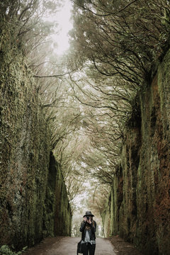 Elegant lady in hat and leather jacket shooting on camera and standing on footpath between murk alley of high walls and woods