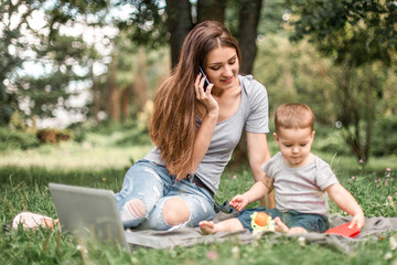 Mother with son sitting in the park, using phone and work on laptop.