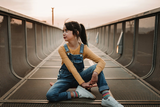 Stylish teen in jean overall posing and sitting on skateboard on footpath in city