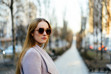 Beautiful young caucasian woman walking along the street in the city. Stylish female model wearing sunglasses walking on city street.