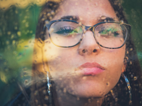 Beautiful young confident lady in eyeglasses and earrings looking at camera through window with water drops