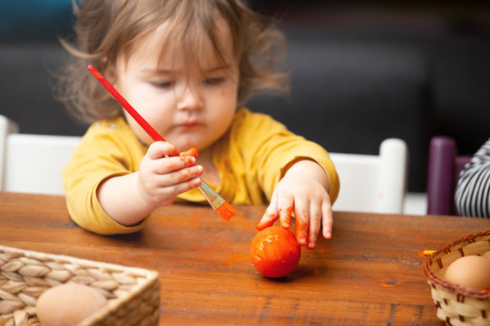 Naklejki Adorable toddler girl using paintbrush to paint Easter egg while sitting at table