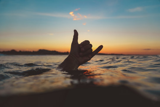 Closeup hand of human showing shaka sign above water surface with ripple and blue heaven in evening on Bali, Indonesia
