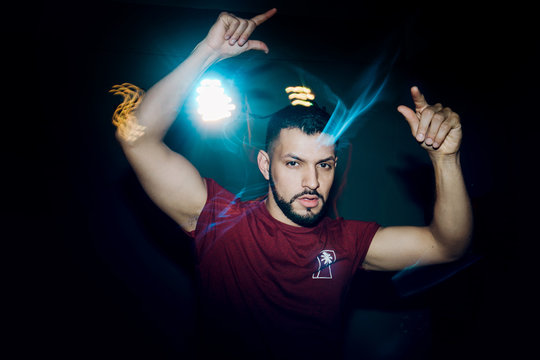 Young handsome man in casual t-shirt holding hands raised and dancing in bright spotlight looking at camera