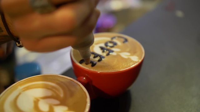 Two red cups of coffee on the table. Barista writes chocolate on the surface of the drink in one of the cups. Writes the word coffee. With one hand holding the other hand. Close-up.