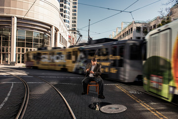 sitting in a chair in the middle of the street downtown reeling in a fish from the sewer with a light rail train in the background.