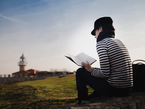 Back view of elegant lady in cap reading volume and sitting on meadow near beacon on shore and wonderful sky
