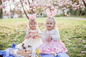 Little cute girls are sitting on the grass near the mogolia. Girls in costumes Easter bunnies.