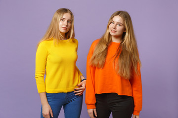 Two young blonde twins sisters girls in vivid colorful clothes standing, looking camera isolated on pastel violet blue wall background in studio. People family lifestyle concept. Mock up copy space.