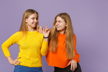 Two smiling young blonde twins sisters girls in colorful clothes looking at each other pointing index finger up isolated on violet blue background. People family lifestyle concept. Mock up copy space.