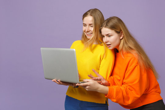 Two smiling young blonde twins sisters girls in vivid colorful clothes holding, using laptop pc computer isolated on pastel violet blue background. People family lifestyle concept. Mock up copy space.