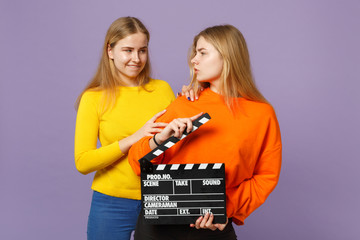 Two funny young blonde twins sisters girls in colorful clothes holding classic black film making clapperboard isolated on violet blue background. People family lifestyle concept. Mock up copy space.