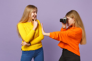 Two young blonde twins sisters girls in vivid colorful clothes taking pictures on retro vintage photo camera isolated on violet blue background. People family lifestyle concept. Mock up copy space.