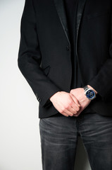 Cropped head man in black blazer holding his hands on the front with stainless steel watch on his right hand, grey wall background