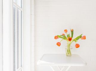 Orange tulips in glass vase on white table against painted brick wall next to window