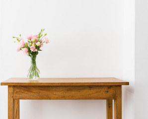 Pink and cream lisianthus flowers in glass vase on oak wooden sidetable against white wall