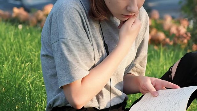 close-up teen girl in a gray t-shirt sits on the grass in the park looks around and reads a notebook with lecture notes on a sunny day