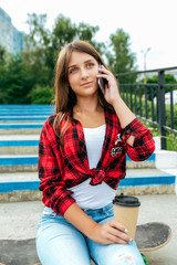 Girl teenager sits on a skateboard, talking on the phone, holding a cup with tea in her hands. In the summer in the city in nature.