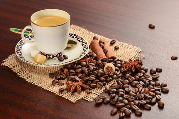 Coffee in porcelain cup on saucer with cinnamon, nutmeg and star anise on handmade jute napkin. Vintage coffee set on wood table with scattered coffee beans.