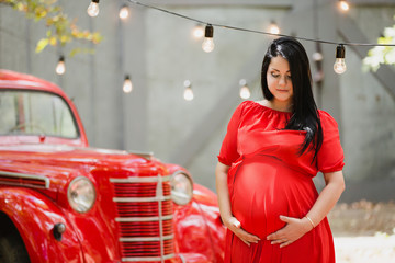 Young pregnant woman in a autumn garden near red retro car. Caucasian woman with long black hairs in long romantic red dress.  Waiting for baby. 