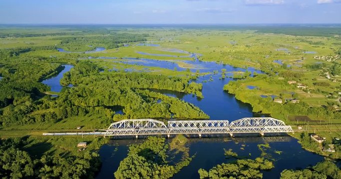 Aerial view of electric train on the railway bridge on the background of beautiful Desna river and flooded meadow at spring. Amazing water landscape of a flood-meadow. White iron bridge at countryside