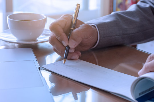 Business man hand writing and signing checkbook with laptop computer and white cup of coffee on the wooden table at modern home office.	