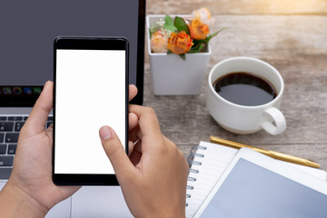 Mock up image of female hand holding and using black mobile smart phone with blank white screen on laptop computer background with tablet and cup of coffee on wooden table at home . clipping path.