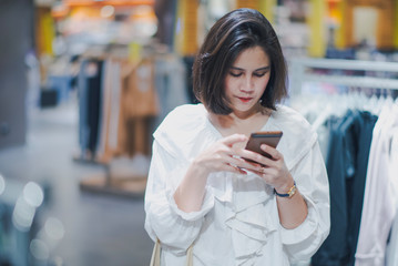 Young Asian beautiful woman shot hair shopping new clothes in a shopping mall store. Woman standing and holding a shopping bag and looking to her mobile phone in the store.