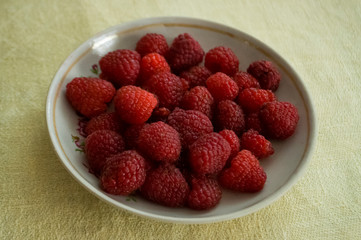 Ripe raspberries are laying on the white plate which is standing on the table covered with yellow tablecloth