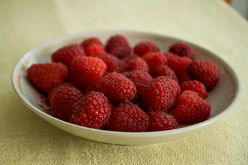 Ripe raspberries are laying on the white plate which is standing on the table covered with yellow tablecloth
