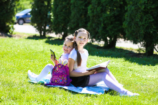 two schoolgirls sit on the lawn in the open air with a book in their hands